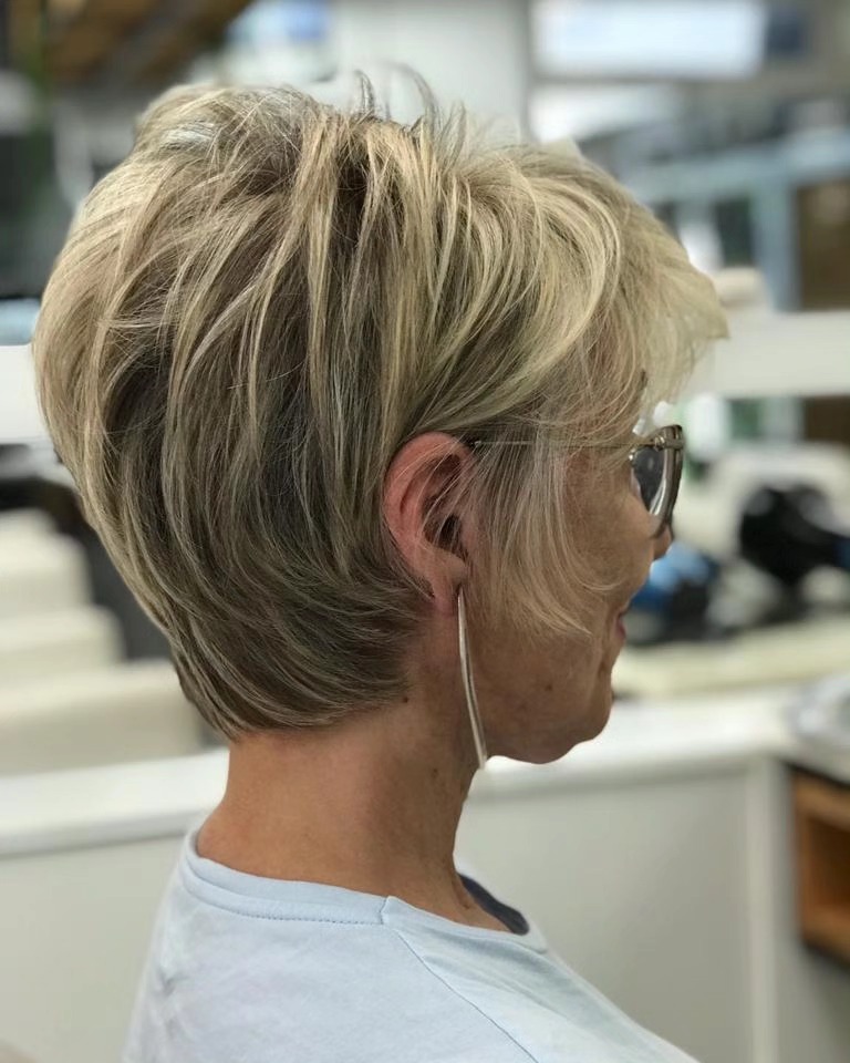 A woman over 60 with a voluminous, feathered beige-blonde pixie bob featuring high lift at the crown and wispy side layers.