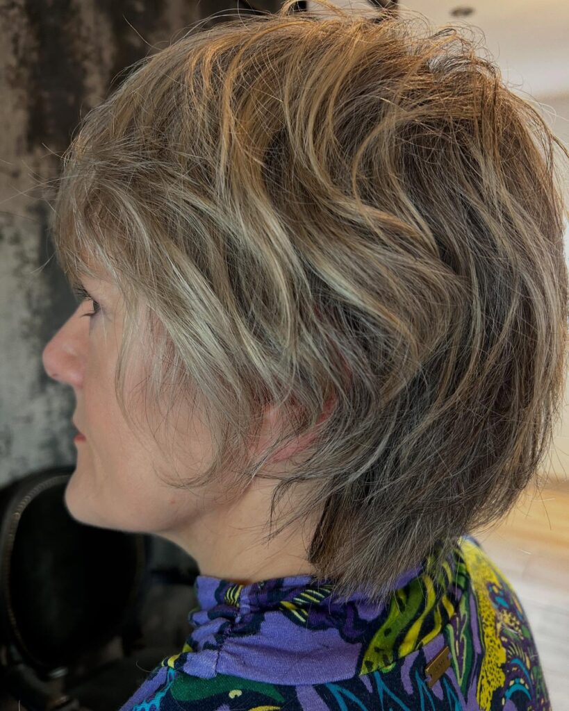A side profile of a woman with a piecey sandy blonde bixie cut featuring choppy layers and a dark shadow root.