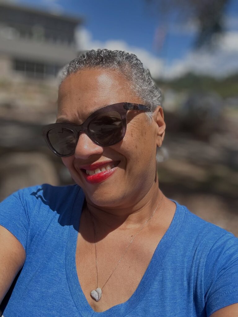 A woman with dark cat-eye sunglasses smiles while sporting a short tapered natural hairstyle with grey curls.