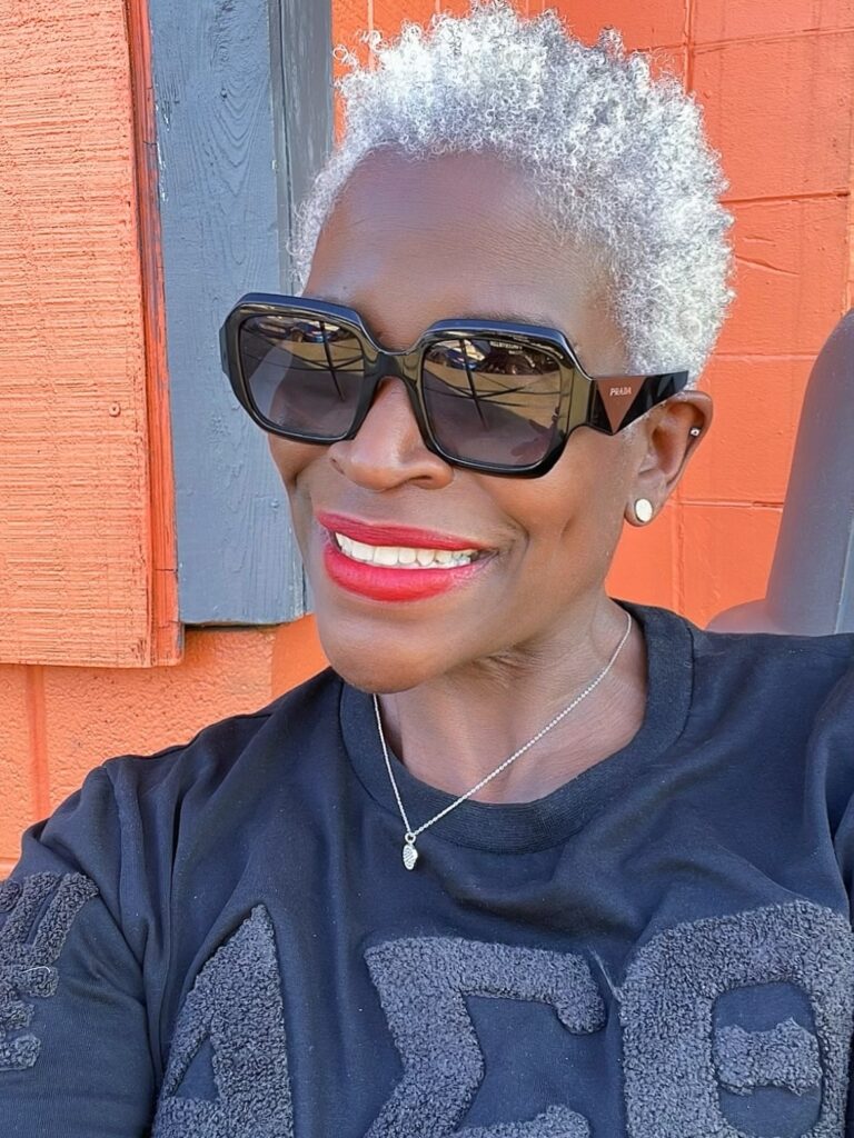 A woman wearing large black sunglasses smiles while showing off a short silver natural hairstyle with tight coils.