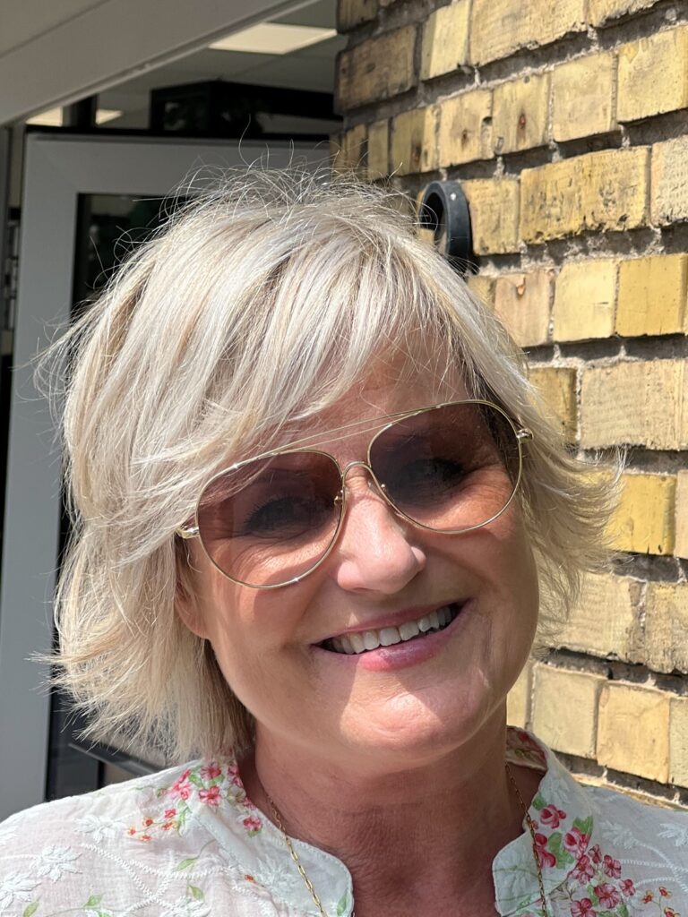 A woman with gold aviator sunglasses smiles while wearing a light champagne-blonde pixie cut with feathery layers and flared ends.