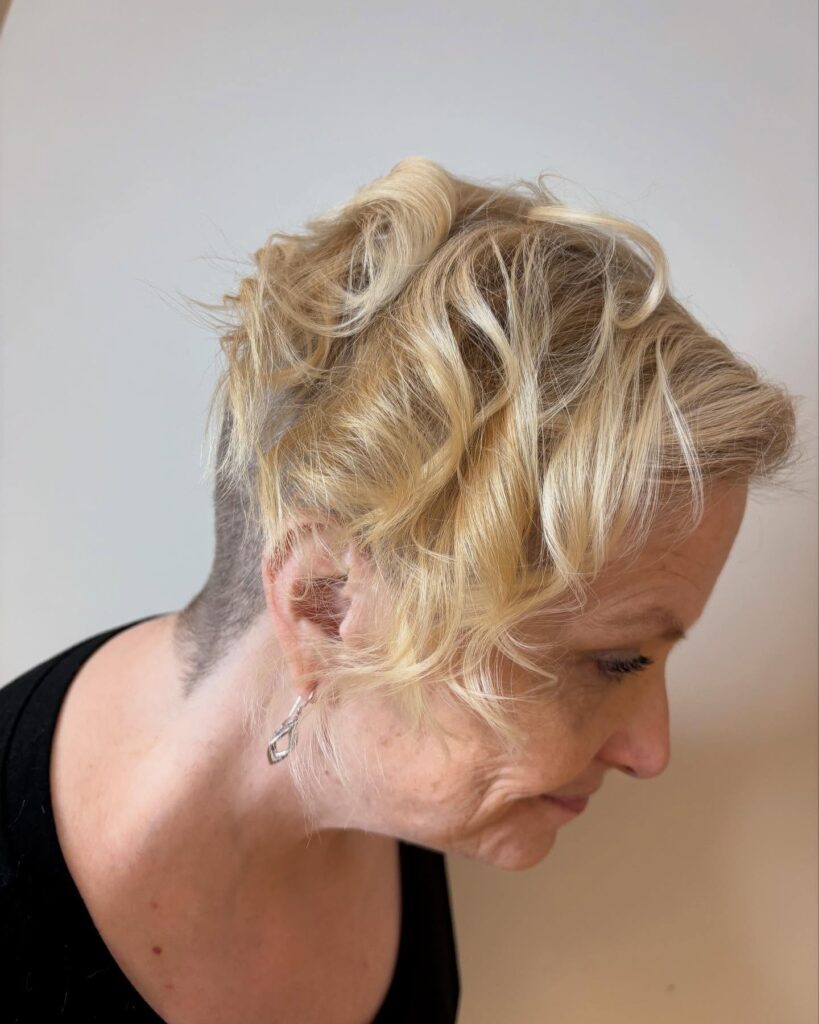 A woman over 60 with blonde curly hair on top and a closely shaved undercut on the side and back.