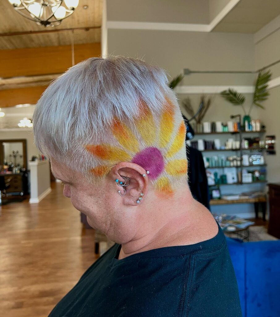 A woman with short silver hair featuring a side undercut, shaved and dyed with a colorful sunflower design in yellow and purple.