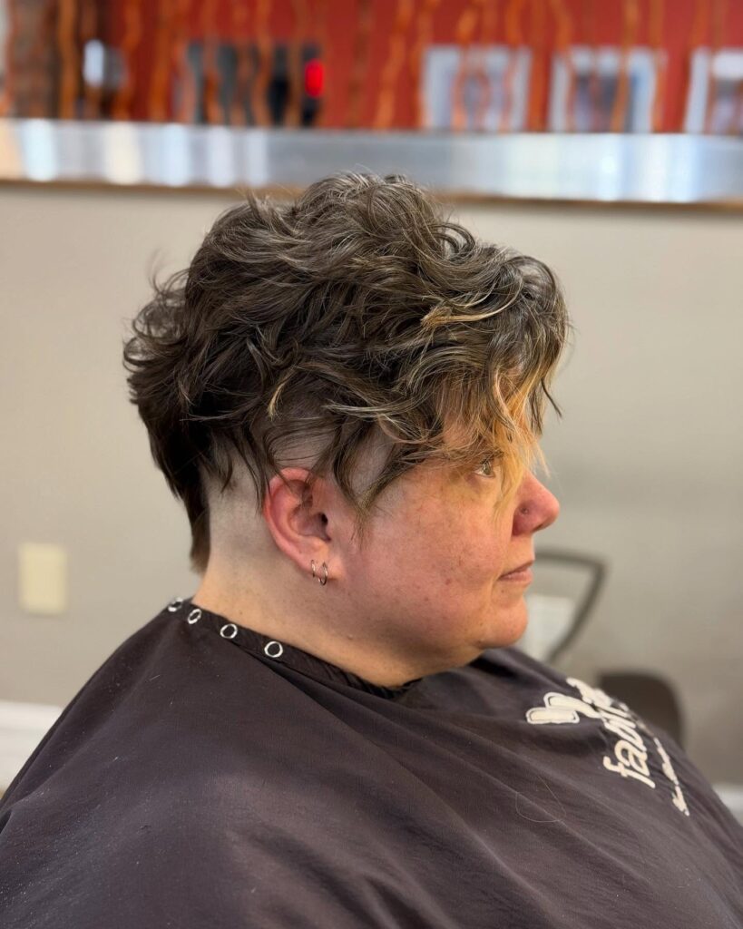 A woman with wavy brunette hair and caramel highlights, featuring a high skin fade undercut around her ear.