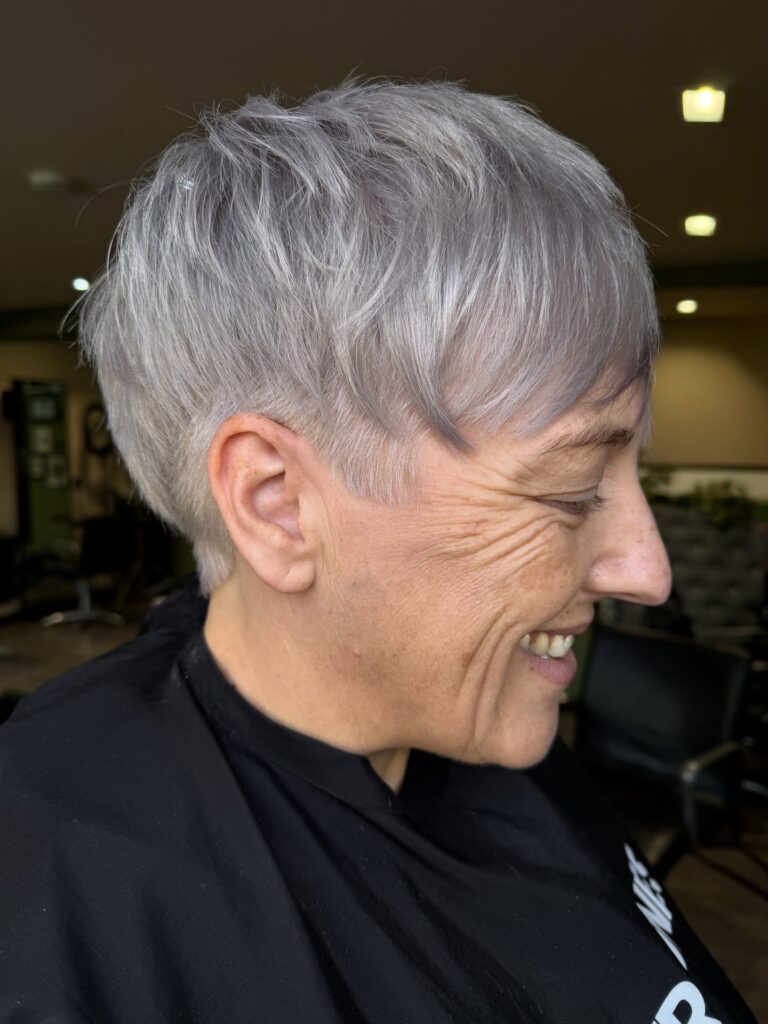 A smiling woman over 60 with a textured smoky silver pixie cut featuring a shaved undercut at the temples.