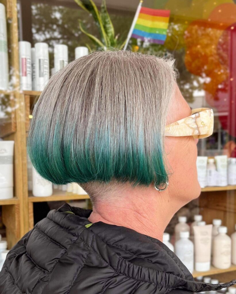 A woman with a straight silver bob featuring bright teal green ends and a hidden shaved undercut at the nape of the neck.
