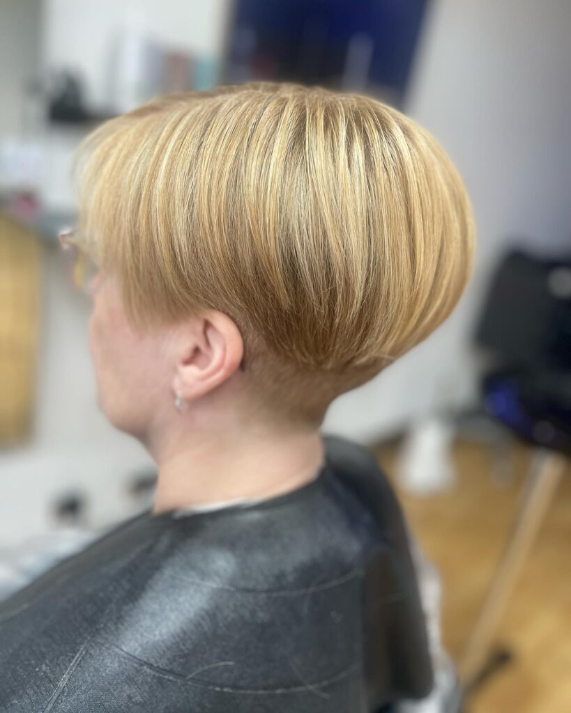 A woman in profile with a structured pixie cut and a honey blonde hair color featuring smooth, long layers on top and a close, tapered shave on the sides and neck.