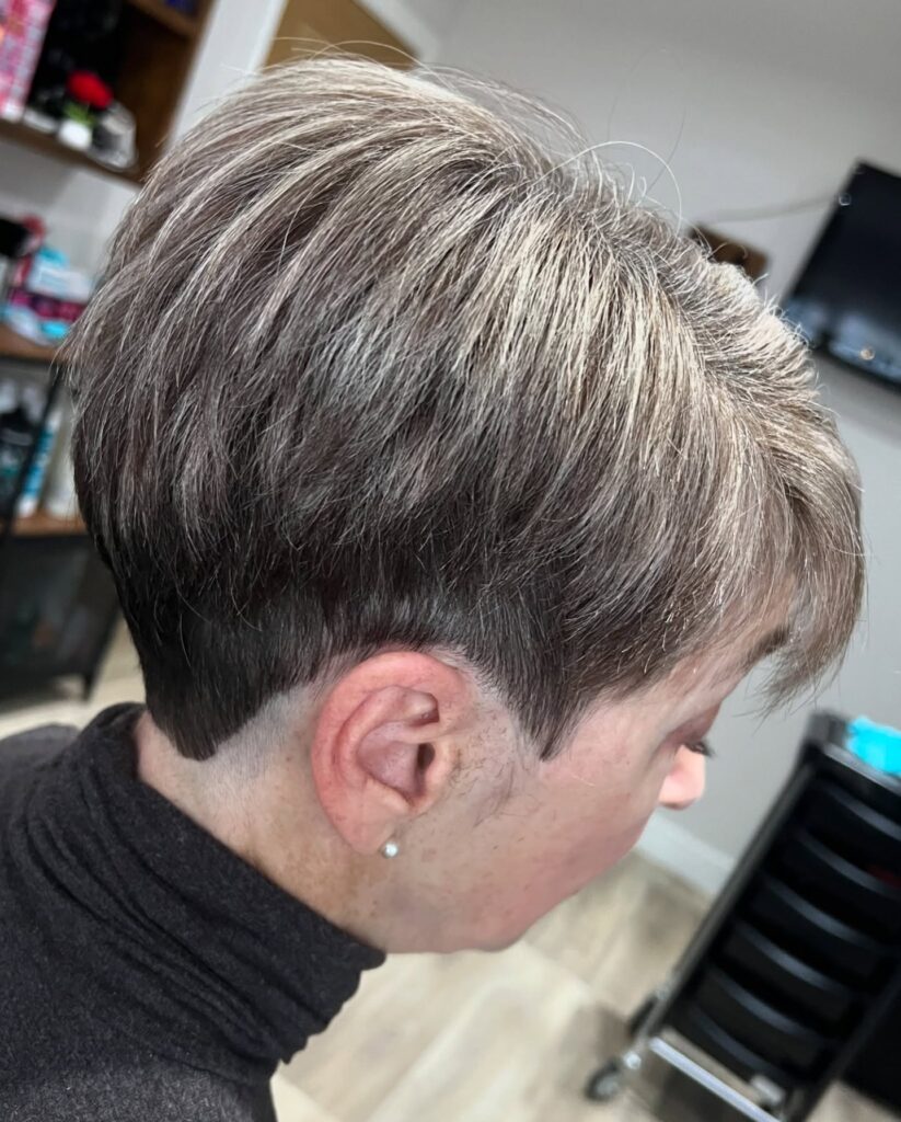 A woman over 60 with a straight ash brown and silver pixie cut featuring a sharp V-shaped buzzed undercut at the nape.