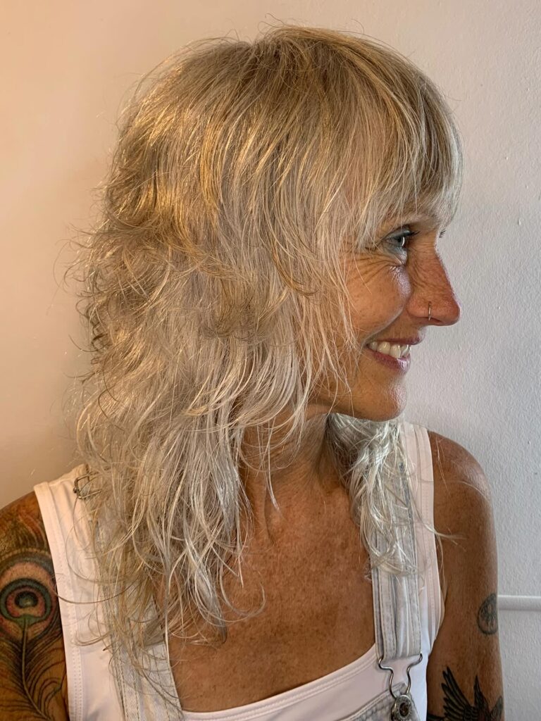 A smiling woman over sixty with a medium-length ash blonde shag haircut featuring a full fringe and voluminous crown layers.