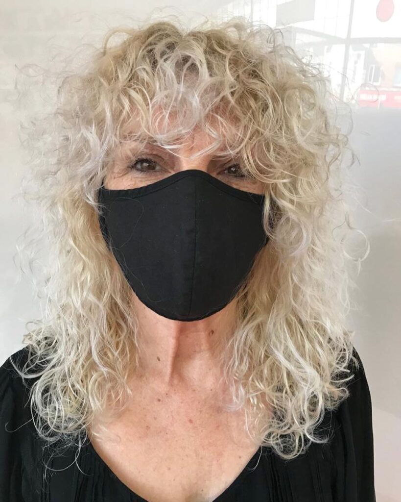 A woman with shoulder-length platinum blonde curly hair styled in a layered shag with a full curly fringe.