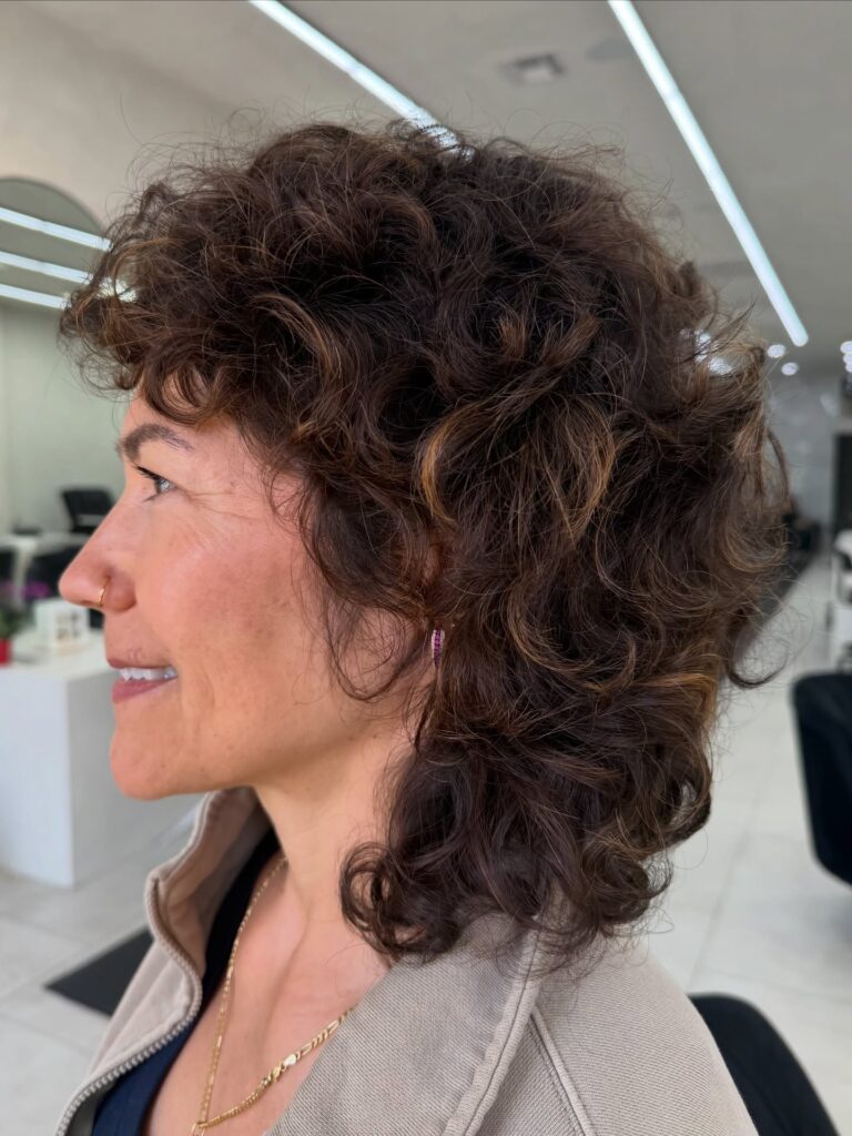 A woman over sixty with a dark chocolate brown curly shag haircut featuring honey highlights and voluminous, rounded layers.