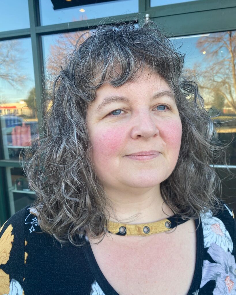 A woman with shoulder-length salt-and-pepper wavy hair styled into a textured shag with piecey bangs.
