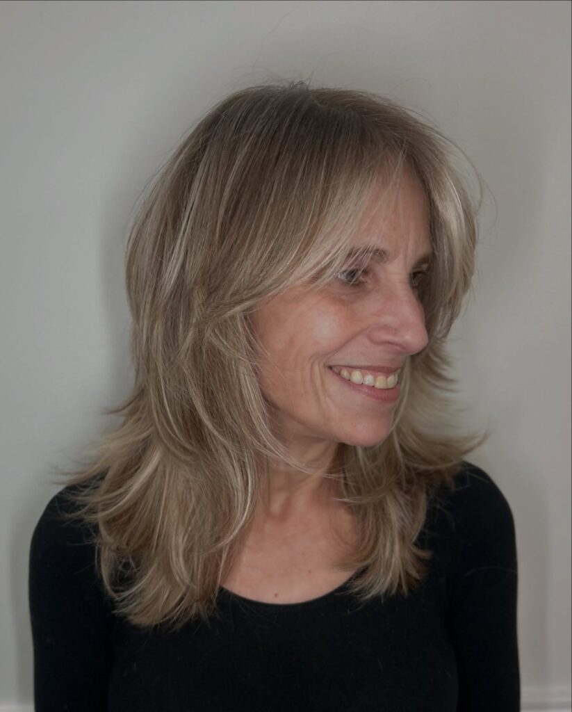 A woman over sixty with a shoulder-length sandy blonde shag haircut featuring feathered layers and long curtain bangs.
