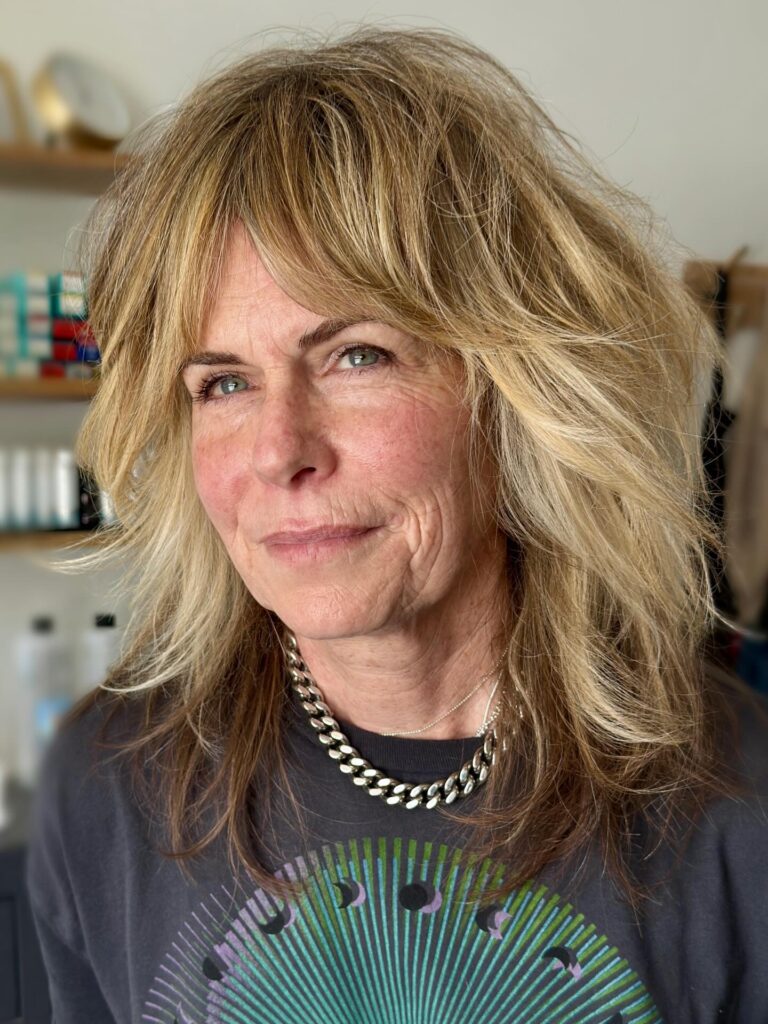 A woman with a medium-length honey blonde shag hairstyle featuring choppy layers and full, piecey bangs.