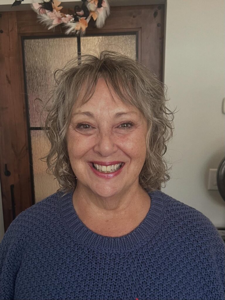 A smiling woman with a shoulder-length wavy mousy brown shag hairstyle and thin wispy bangs.
