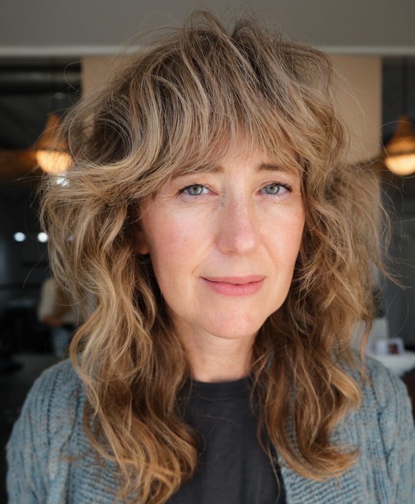 A woman with shoulder-length sandy-brown wavy hair in a shaggy layered cut with sweeping curtain bangs.