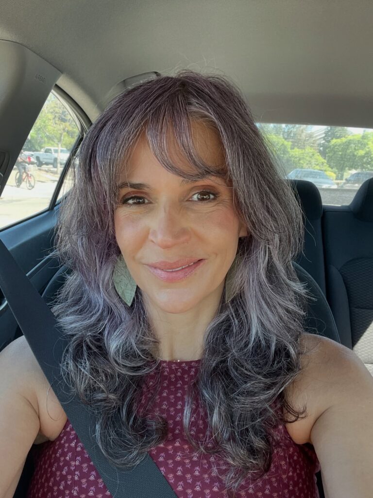 A woman over sixty with a long shag haircut featuring lavender and silver tones, a wispy fringe, and wavy layers.
