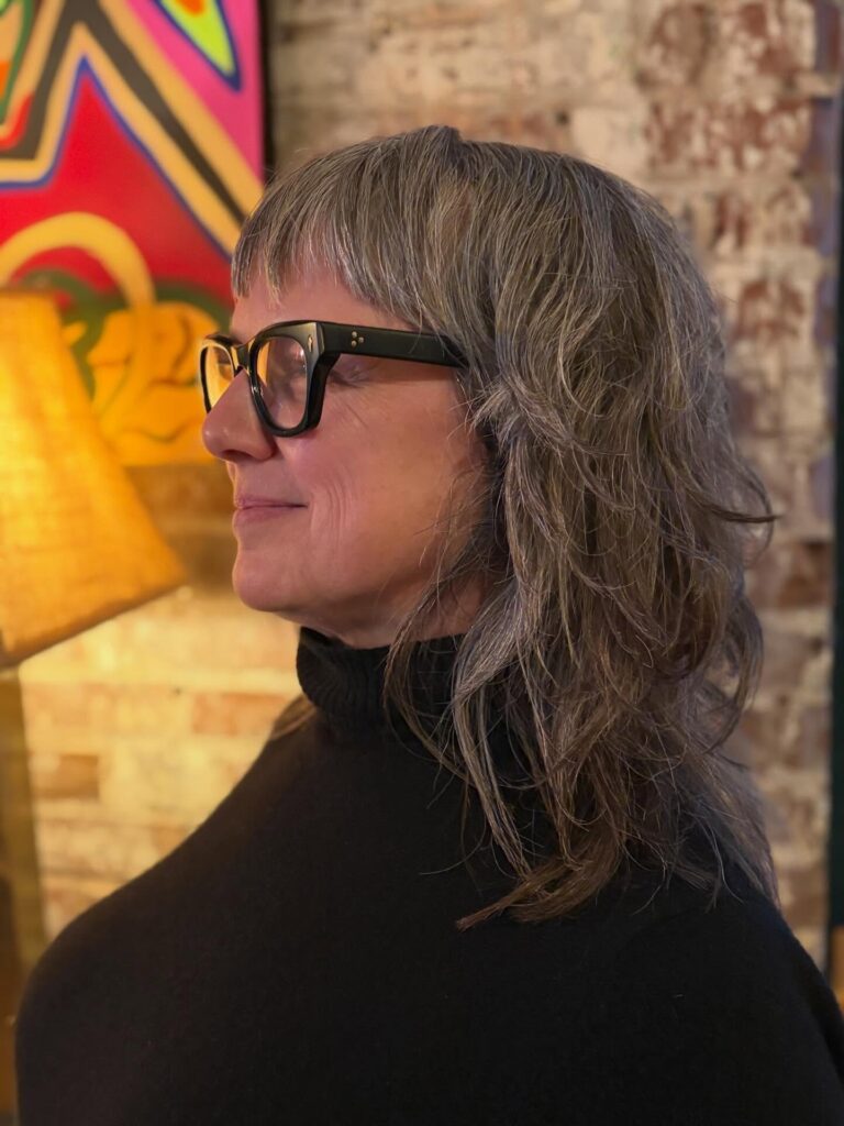 A woman over sixty with a medium-length smoky grey shag haircut featuring a short micro fringe and wispy layers.