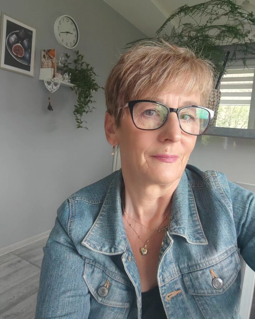 A woman with a sandy blonde tapered pixie cut featuring soft layered bangs and wearing black-framed glasses.