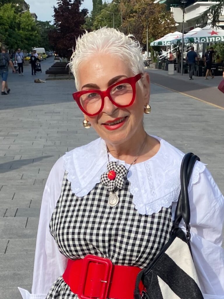 A stylish woman over 60 with a short, spiky, snow-white pixie cut and bright red glasses.