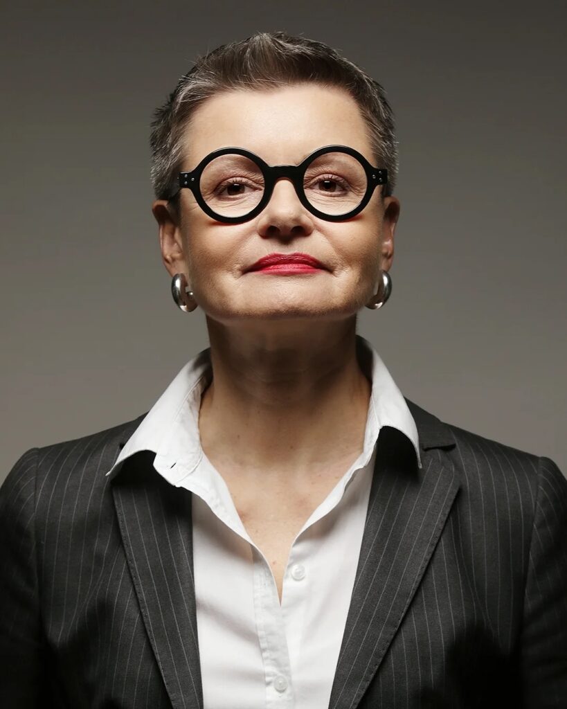 A woman over 60 with a short textured salt and pepper pixie cut and bold round black glasses wearing a pinstripe blazer.