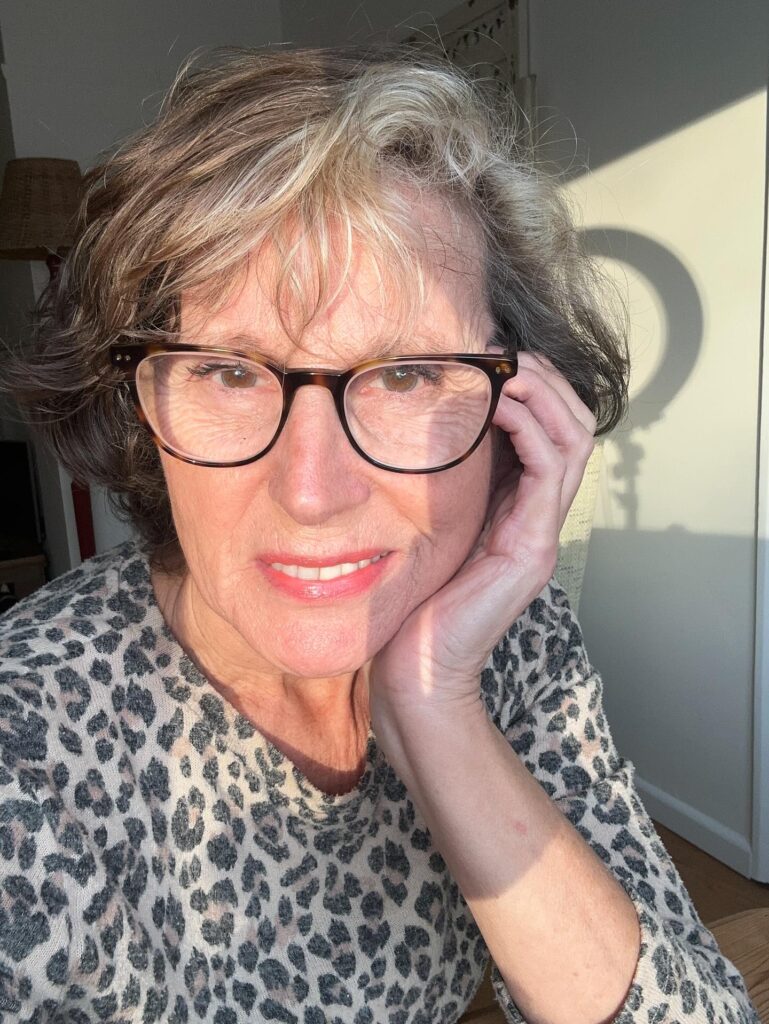 A woman over 60 with a wavy ash brown chin-length bob and wispy bangs, wearing tortoiseshell glasses and a leopard print top.
