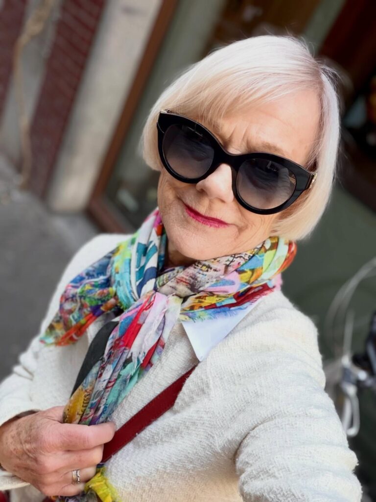 A woman over 60 with a straight platinum blonde jaw-length bob and side-swept bangs wearing large black sunglasses and a colorful scarf.
