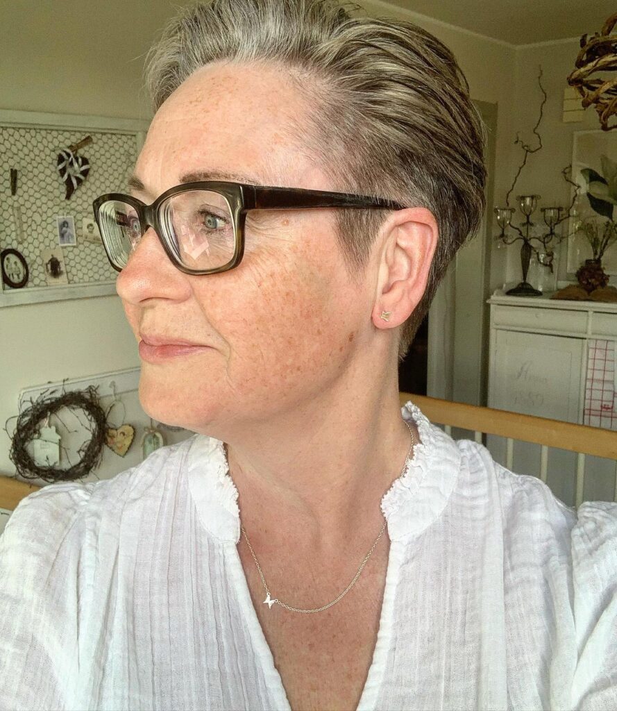 A woman in profile wearing dark-rimmed glasses with a short ash blonde pixie cut that is swept back away from her face.