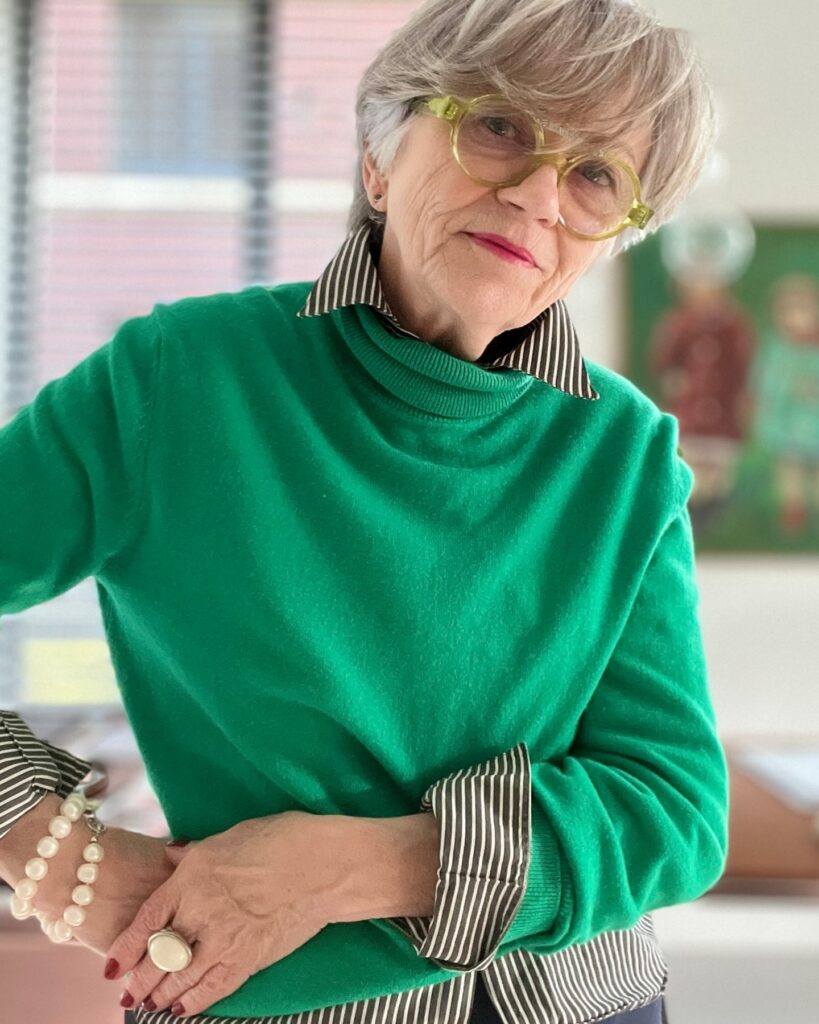 A woman over 60 with a soft ash-blonde pixie cut and textured bangs, wearing translucent lime green glasses and a bright green sweater.
