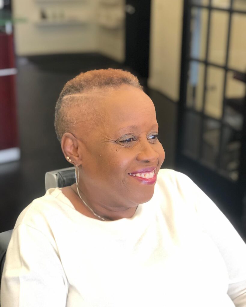 A smiling older Black woman with a honey brown buzzed haircut featuring a sharply shaved side part.