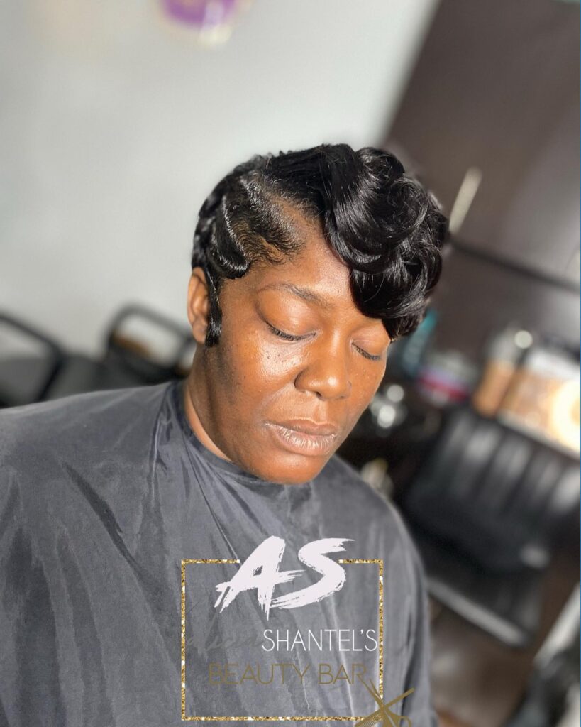 A Black woman wearing a sophisticated hairstyle featuring sleek finger waves on the sides and large, polished curls on top.