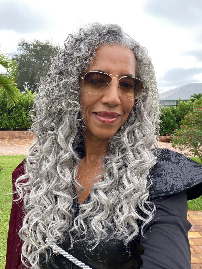 A woman with long cascading silver curls and aviator sunglasses is smiling outdoors.