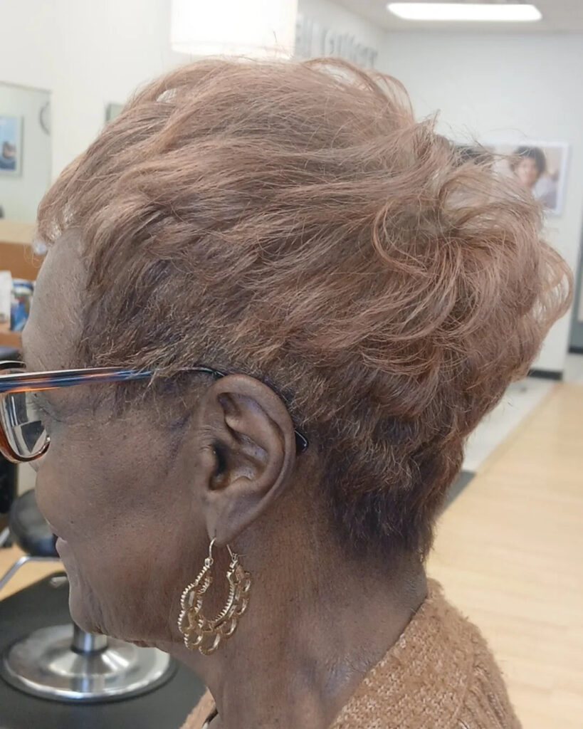 A profile view of an older Black woman with a textured auburn pixie cut featuring wispy layers and a tapered back.