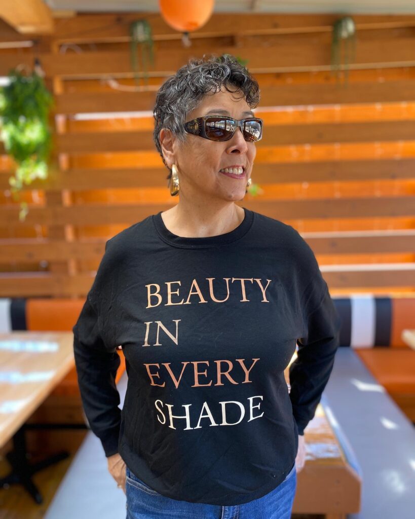 An older woman with a short charcoal grey curly pixie cut is smiling while wearing sunglasses.
