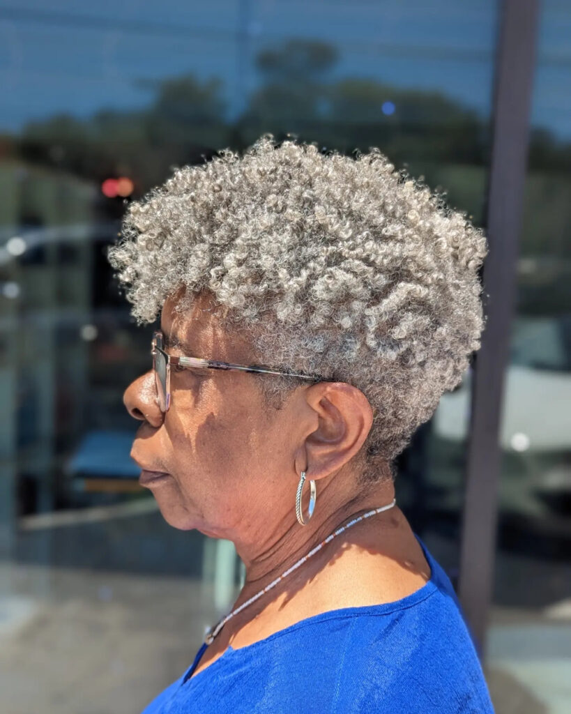 A profile view of an older Black woman featuring a voluminous and rounded silver natural curly hairstyle.