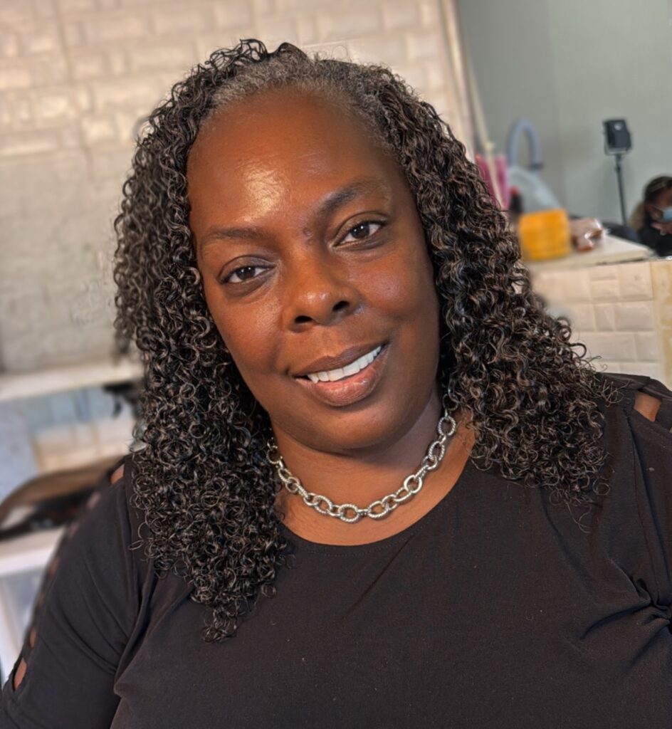 A Black woman over 60 with shoulder-length defined natural coils in a salt-and-pepper color styled with a middle part.