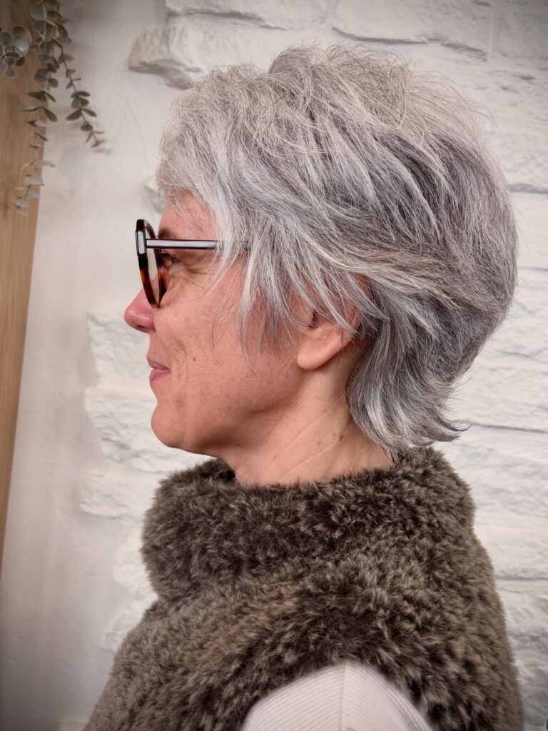 A woman with short layered grey hair and stylish glasses is showing a side profile of her textured pixie cut.