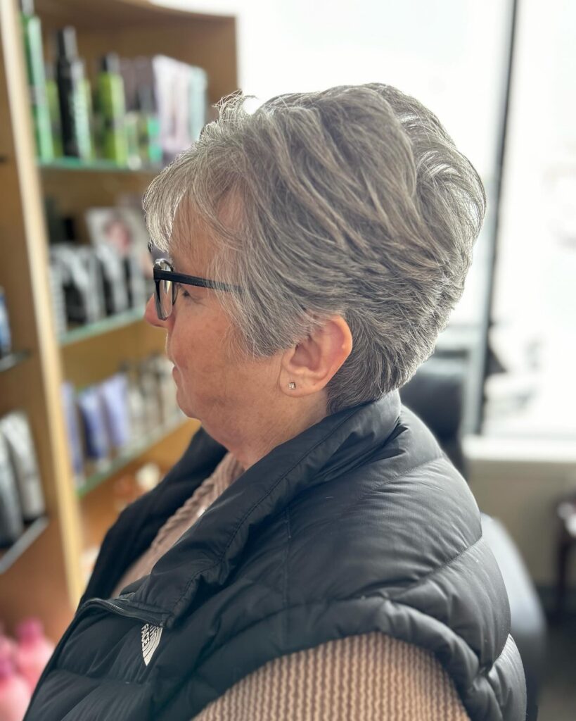 A side view of a woman with a short, spiky, salt-and-pepper pixie cut wearing black glasses.