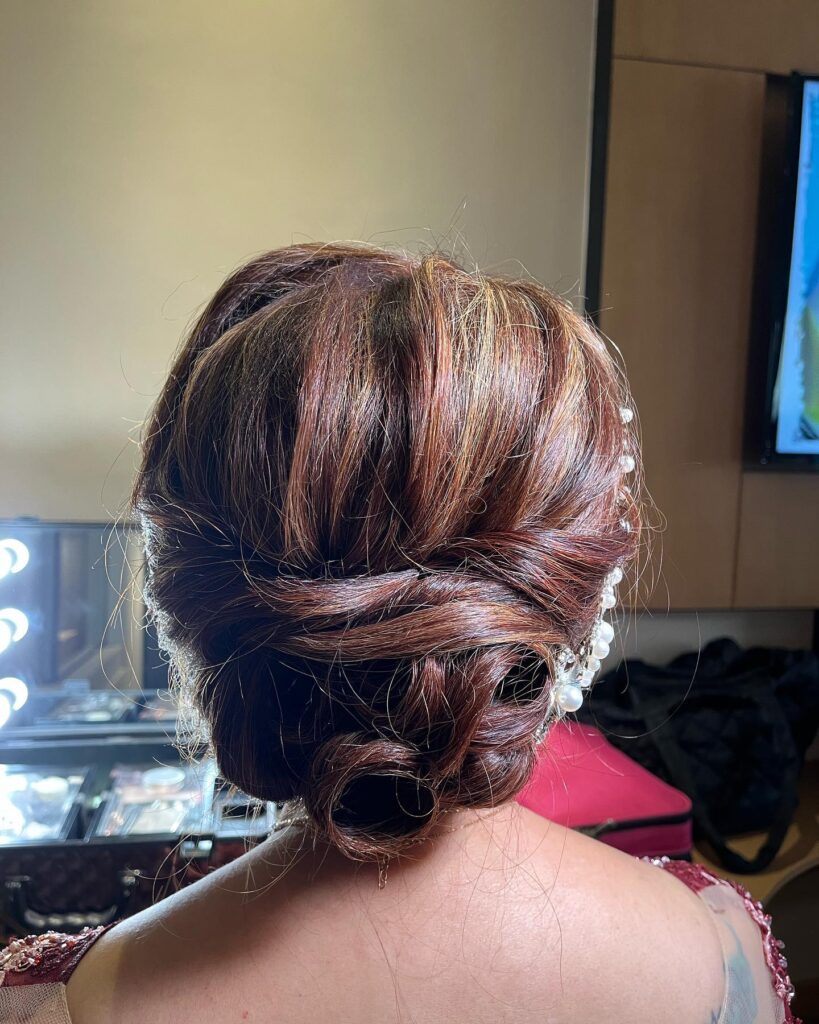 A back view of a woman with auburn hair styled into an intricate low bun accented with a delicate vine of white pearls.