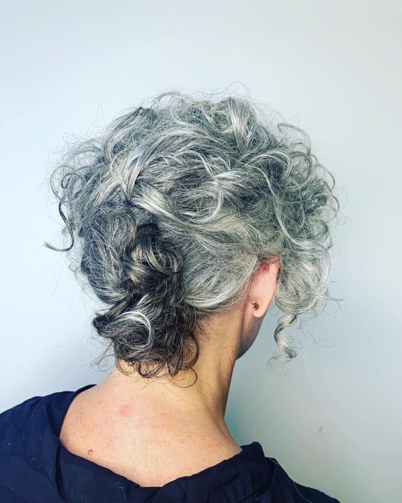 A close-up from behind of a woman with naturally curly salt-and-pepper hair styled into a soft, textured updo.