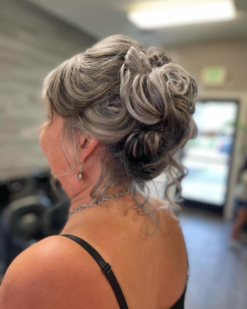 A woman with silver and lilac hair styled into a high-textured updo with decorative loops and loose curls at the neck.