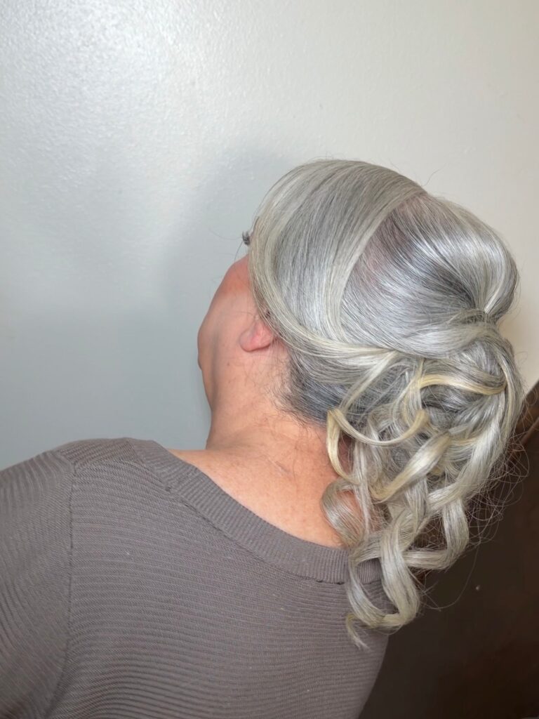 A side view of a woman with silver and blonde hair styled into a sleek updo that transitions into a cascade of defined curls at the back.