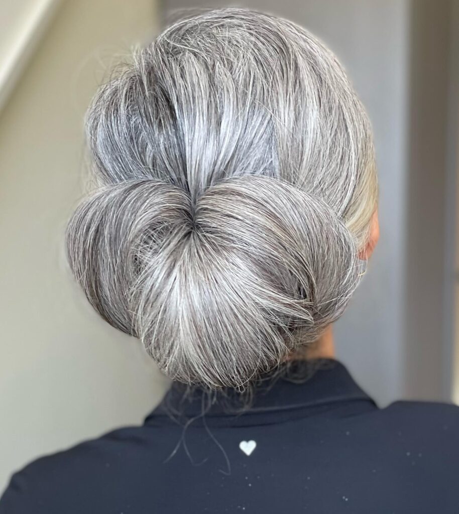 A back view of a woman with thick silver hair styled into a large, sculptural low bun with a unique folded petal shape.