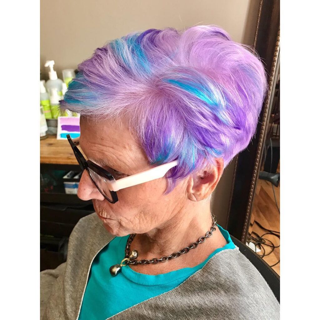 A woman with a short layered pixie cut featuring a mix of lavender, purple, and bright teal hair colors.
