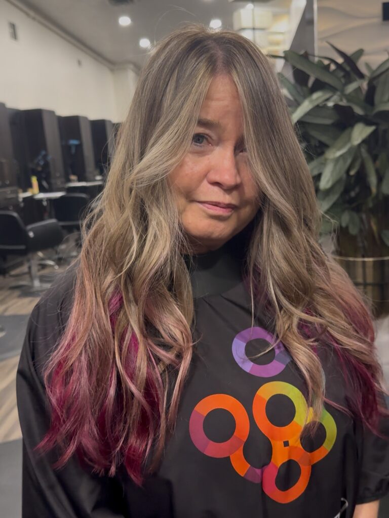 A woman with long, wavy, sandy blonde hair featuring vibrant magenta peekaboo highlights at the ends.