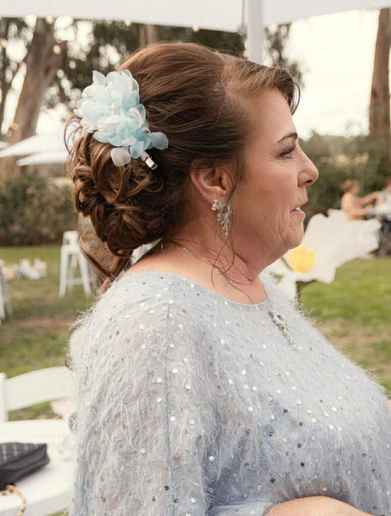 A woman with a curly brunette low bun updo accented by a large light blue flower hair clip.