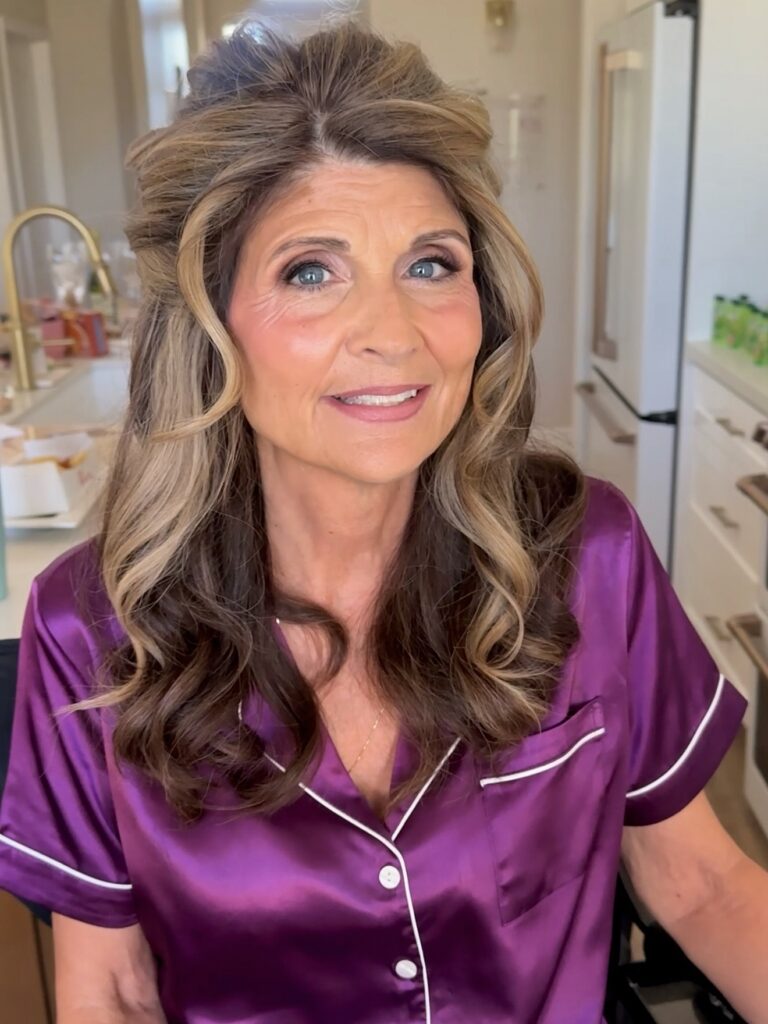 A woman with long, brown, and blonde highlighted hair styled in a voluminous half-up look with large curls.