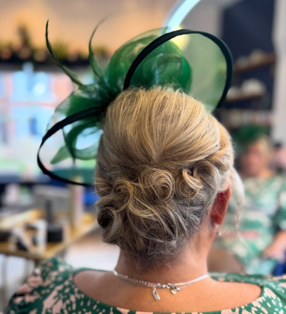 A woman from behind with a blonde curly updo accented by a large green mesh fascinator.