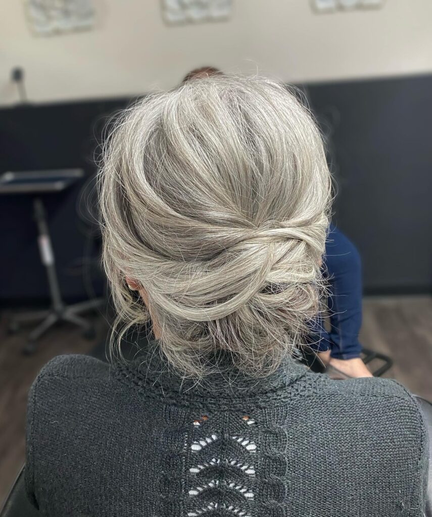 A woman over 60 with salt-and-pepper hair styled into a voluminous low chignon with textured overlapping layers at the back.