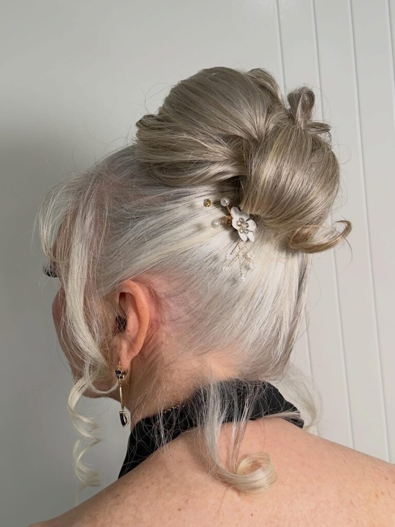 A woman over 60 with bright silver hair styled into a high swirled bun, featuring a pearl floral hairpin and soft face-framing curls.
